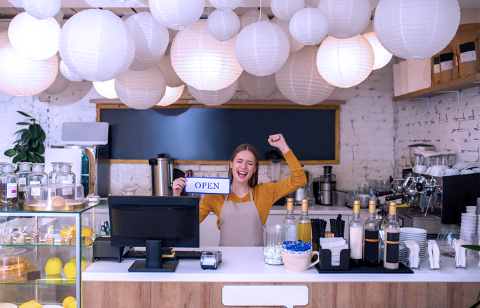 New Small Business Cafe Owner Holding an Open Sign at the Counter Next to Coffee and Cake Displays