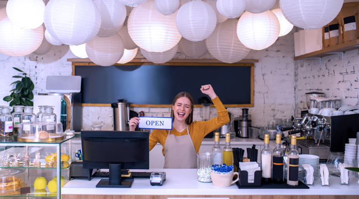 New Small Business Cafe Owner Holding an Open Sign at the Counter Next to Coffee and Cake Displays