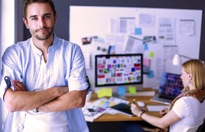Small Business Owners Discussing Marketing Challenges as the Man is Leaning on an Office Wall with a Woman Sitting at a Desk Crunching Numbers