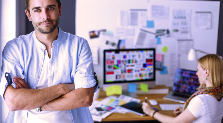 Small Business Owners Discussing Marketing Challenges as the Man is Leaning on an Office Wall with a Woman Sitting at a Desk Crunching Numbers